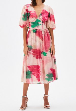 Woman wearing a pink midi dress with green and red abstract floral patterns and puffed sleeves, paired with white high-heeled sandals.