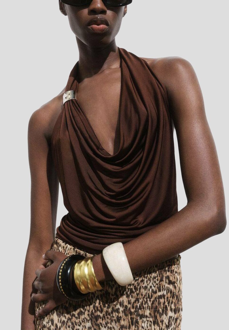 Hnedý halter top zo slabého materiálu s ozdobami z strieborného kovu. Skombinovaný s vzorovanou sukňou a viacerými náramkami z rôznych materiálov.