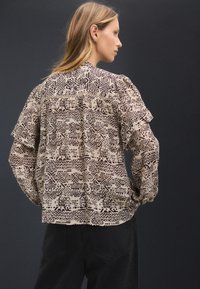Lightweight, patterned blouse in beige and brown, featuring puffed sleeves and a pleated back. Abstract designs cover the fabric.