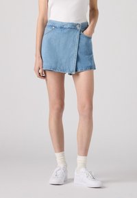 Mini falda de mezclilla en azul claro con diseño  envolvente, bolsillo delantero y cierre de botón en la cintura. Combinada con zapatillas blancas y calcetines.