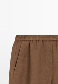 Pantalon marron avec taille élastique et poches latérales, en tissu texturé, présenté de la taille vers le bas sur fond blanc.
