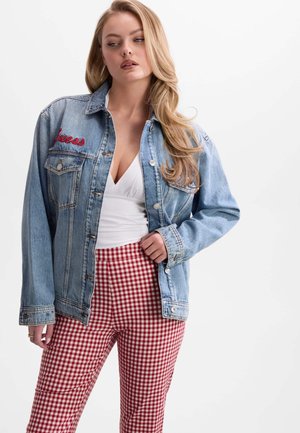 Femme aux longs cheveux blonds portant une veste en jean bleue par-dessus un haut blanc et un pantalon à carreaux rouge et blanc, debout devant un fond uni.