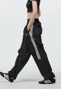 Svarta träningbyxor har tre vita ränder längs sidorna, en avslappnad passform och sidofickor. I kombination med svarta skor och en crop top.