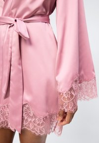 Ρόμπα από ροζ σατέν με ζώνη στη μέση και δαντέλα στα μανίκια και το τελείωμα, εμφανίζεται στον κορμό και το χέρι ενός ατόμου.