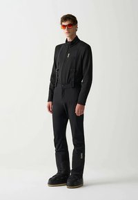 Haut noir zippé associé à un pantalon isolant noir avec bretelles réglables. Accessoires comprennent des lunettes de soleil à verres teintés orange et des chaussures noires.
