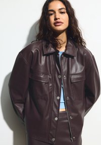Femme aux longs cheveux bouclés portant une veste en cuir marron foncé et un pantalon assorti sur un haut bleu clair, debout devant un fond blanc uni.