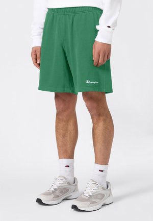 Mann trägt grüne Champion-Shorts, weißes Langarmshirt, weiße Socken und weiße Sneakers, steht vor einem schlichten weißen Hintergrund.