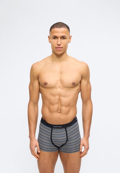 Άνδρας σε φόρμα με κοντά μαλλιά, φορώντας γκρι ριγέ ανδρικά εσώρουχα boxer με μαύρη μέση, στέκεται μπροστά σε απλό ανοιχτό φόντο.