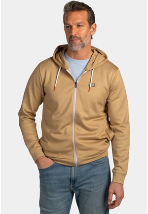 Man draagt een beige hoodie met ritssluiting en witte trekkoorden over een lichtblauw shirt en blauwe spijkerbroek, staand met één hand in zijn zak.