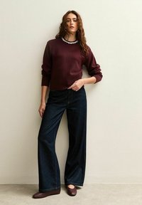 Maglione bordeaux con orlo a coste, pantaloni in denim scuro ampi e ballerine marroni. Il maglione presenta un collo decorativo. Texture lisce in tutto il look.