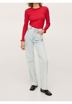 Femme portant un haut rouge ajusté à manches longues avec des bords à volants, un jean taille haute bleu clair et des mocassins noirs, debout avec une main dans la poche.