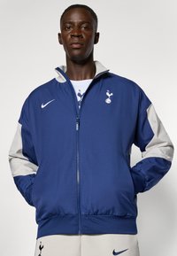 Giacca zip-up navy con maniche grigio chiaro, caratterizzata da un logo sul petto. Realizzata in un tessuto morbido con polsini e vita elasticizzati.