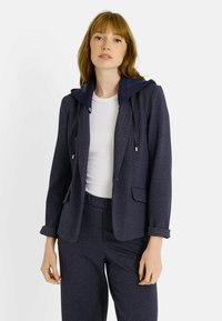 Blazer azul marino con capucha, confeccionado en una tela texturizada con un patrón sutil. Estilizado con una blusa blanca y pantalones azul marino a juego.