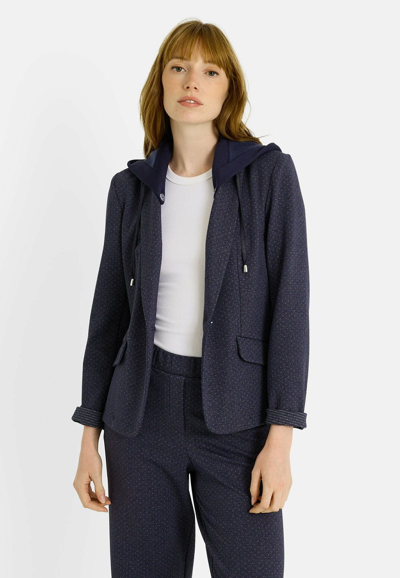 Blazer azul marino con capucha, confeccionado en una tela texturizada con un patrón sutil. Estilizado con una blusa blanca y pantalones azul marino a juego.