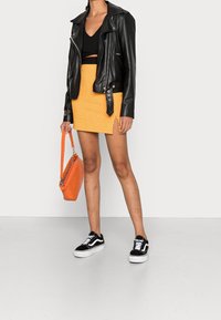 Una mujer está de pie con una chaqueta de cuero negra, una blusa negra y una falda corta naranja, sosteniendo un bolso naranja y usando zapatillas negras.