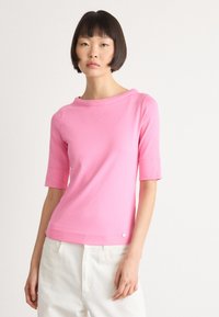 Vrouw met kort donker haar, gekleed in een roze top met halve mouwen en witte broek, tegen een effen witte achtergrond.