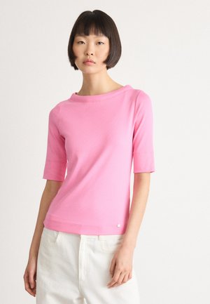 Vrouw met kort donker haar, gekleed in een roze top met halve mouwen en witte broek, tegen een effen witte achtergrond.