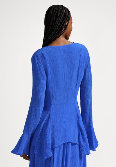 Naine pikkade punutud juustega, seljas tekstuuriga kuninglikult sinine pika varrukaga pluus lehvivate mansettide ja asümmeetrilise äärega, vaadatuna tagant.