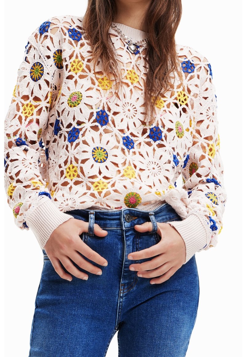 Maglione all'uncinetto in rosa chiaro con motivi floreali colorati in blu, giallo e verde. Presenta polsini e orlo a coste, indossato sopra jeans blu.