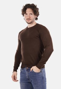 Uomo con capelli ricci e barba che indossa un maglione marrone e jeans blu, in piedi con una mano in tasca su uno sfondo bianco.