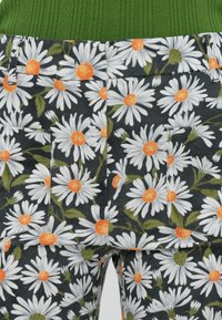 Vue arrière d’un pantalon foncé avec un motif de fleurs de marguerite blanche aux centres orange, porté avec une chemise verte côtelée rentrée.