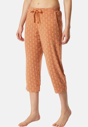 Frau trägt orangefarbene, gekürzte Hose mit weißem Blumenmuster und Kordelzug in der Taille und steht barfuß vor einem einfarbigen Hintergrund.