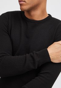 Czarny sweter o fakturze z okrągłym dekoltem i prążkowanymi mankietami. Tkanina ma wzór plastra miodu i dopasowany krój.