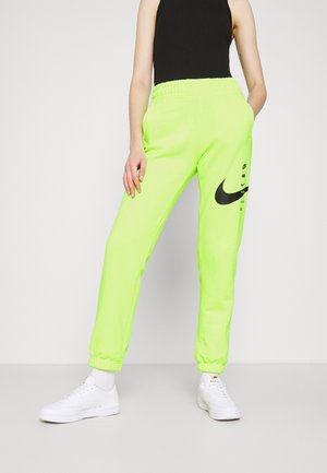 Personne portant un pantalon de survêtement Nike vert néon, un haut sans manches noir et des baskets blanches, debout les mains dans les poches sur un sol gris.