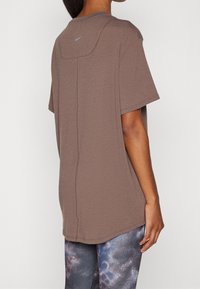 Camiseta marrón de manga corta con cuello redondo, textura suave y un pequeño logo azul en la parte trasera. Combinada con leggings estampados.