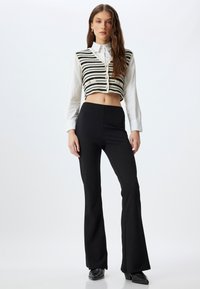 Pantalones acampanados negros de textura suave, combinados con un chaleco de punto a rayas en negro y crema con botones y recorte. Camisa blanca de manga larga debajo.