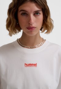 Hvid bomulds t-shirt med rund hals og korte ærmer. Centreret rødt "hummel PERFORMANCE" logo på brystet. Sølvkæde halskæde.