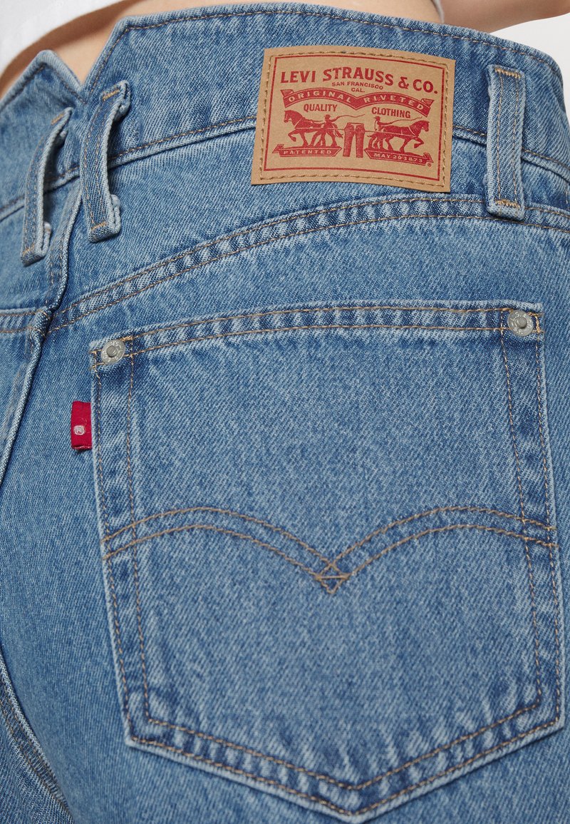 Jeans de denim azul con un parche de cuero beige, etiqueta de logo roja y costuras en contraste. Presentan un diseño clásico de bolsillo trasero con remaches.