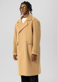 Man met gevlochten haar, gekleed in een oversized dubbelbreasted camel kleurige jas, wit overhemd, zwarte broek en geel getinte bril, tegen een grijze achtergrond.