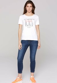 Camiseta blanca de manga corta con un estampado gráfico, combinada con unos vaqueros ajustados azul oscuro y zapatillas naranjas. Conjunto sencillo y casual.