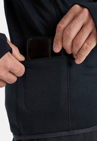 Top nero a maniche lunghe con un design aderente, dotato di una tasca laterale per uno smartphone e di un tessuto testurizzato lungo il corpo.