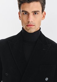 Młody mężczyzna z ciemnymi włosami, ubrany w czarny golf i podwójnie zapiętą czarn�ą marynarkę, patrzy prosto w aparat z neutralnym wyrazem twarzy.