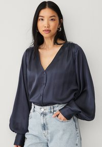 Marine satinbluse med V-udskæring, lange pufærmer og knapper ned foran. Parret med lyse jeans.