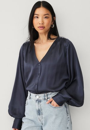 Blusa di satin blu navy con scollatura a V, maniche lunghe a sbuffo e bottoni sul davanti. Abbinata a jeans in denim chiaro.