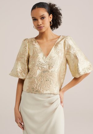 Ženska z kodrastimi lasmi, oblečena v zlato cvetlično brokatno bluzo z V-izrezom in napihnjenimi rokavi ter svetlo bež krilo, stoji z eno roko na boku.