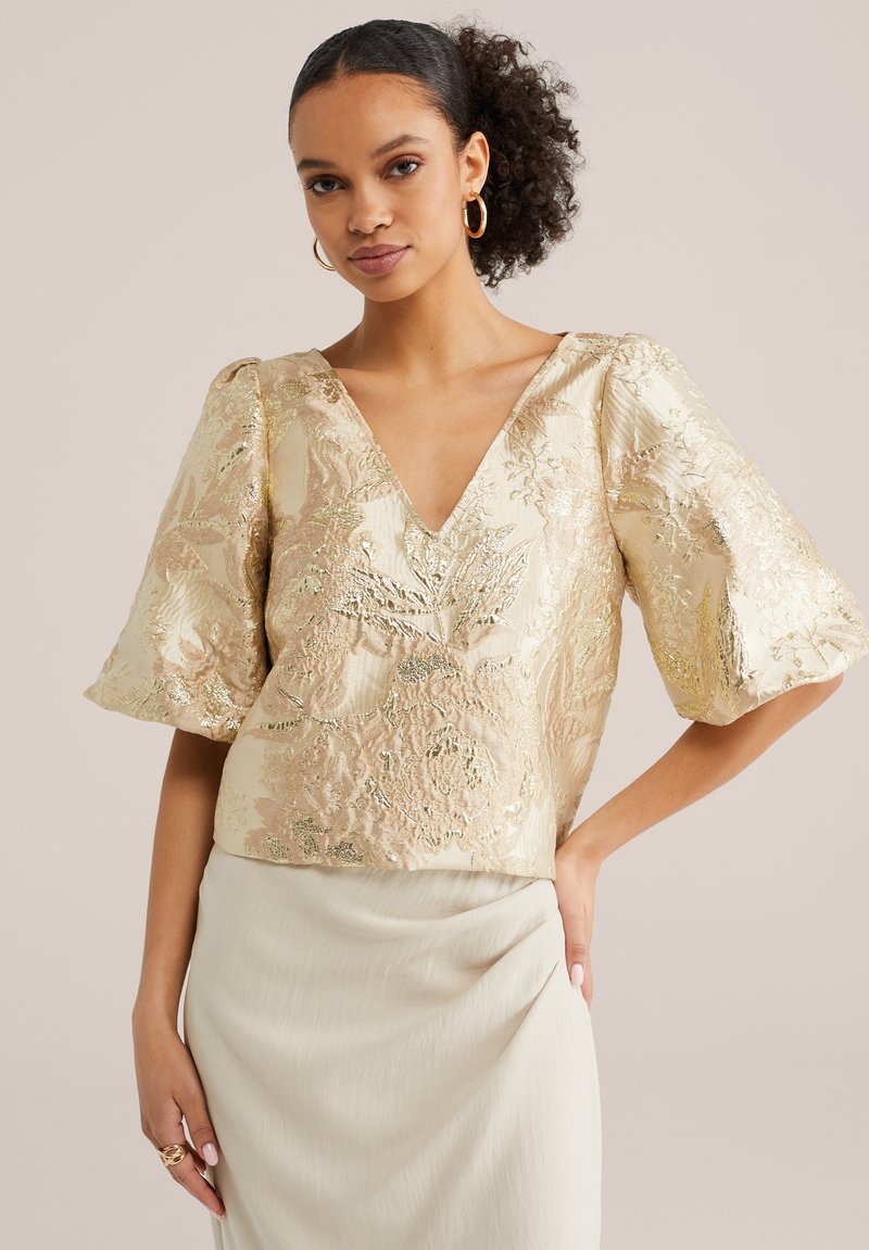 Femme aux cheveux bouclés portant un chemisier en brocart doré à motifs floraux avec col en V et manches bouffantes, associée à une jupe beige clair, debout avec une main sur la hanche.