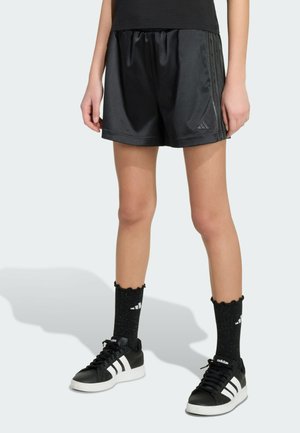 Osoba nosząca czarne sportowe szorty Adidas, czarne skarpetki z białym logo oraz czarne buty sportowe Adidas z białymi paskami, stojąca na jasnym tle.
