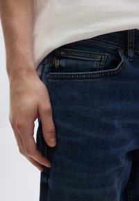 Traperice tamnoplave boje s glatkom teksturom, s vidljivim džepom i etiketom. Ruka se oslanja na tkaninu. Košulja je jednobojna bijela.