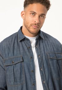 Denim button-up shirt met twee borstzakken, gestikte details en een kraag, gedragen over een wit T-shirt.