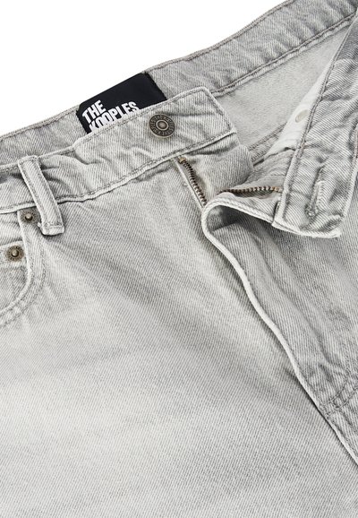 Jeans en denim gris clair avec fermeture éclair partiellement ouverte, bouton de fermeture, rivets sur les poches, et étiquette noire portant l'inscription "THE KOOPLES."