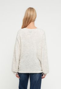 Sweater oversize bege claro com textura suave, punhos canelados e decote largo, combinado com jeans azuis. Vista traseira mostrada.