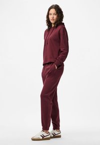 Una mujer se encuentra de pie con las manos en los bolsillos, vistiendo una sudadera burdeos, joggers a juego y zapatillas blancas, frente a un fondo blanco.
