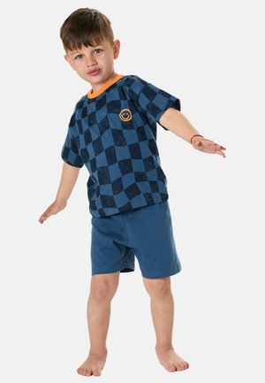 Young boy balancing on one foot wearing blue checkered shirt with smiley face and blue shorts, barefoot on plain background.