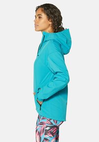 Türkise Softshell-Jacke mit Kapuze, Reißverschlusstaschen und einem eleganten Design. Über gemusterten Leggings mit pinken und grünen Blättern getragen.