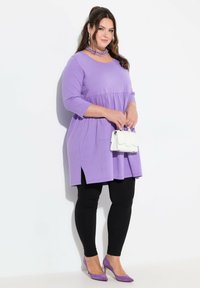 Lila tunika med en utsvängd design, trekvartslånga ärmar, sidoklyftor; kombinerad med svarta leggings och lila klackar, håller en vit handväska.