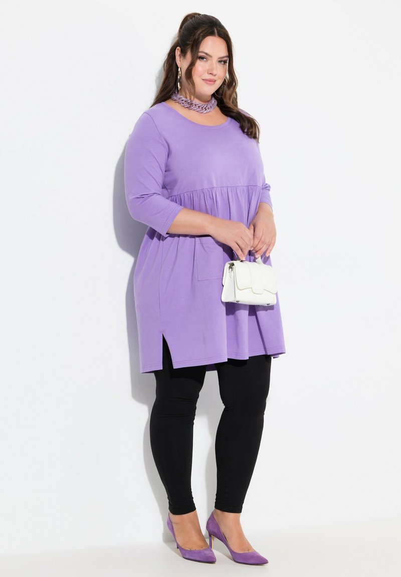 Lila tunika med en utsvängd design, trekvartslånga ärmar, sidoklyftor; kombinerad med svarta leggings och lila klackar, håller en vit handväska.